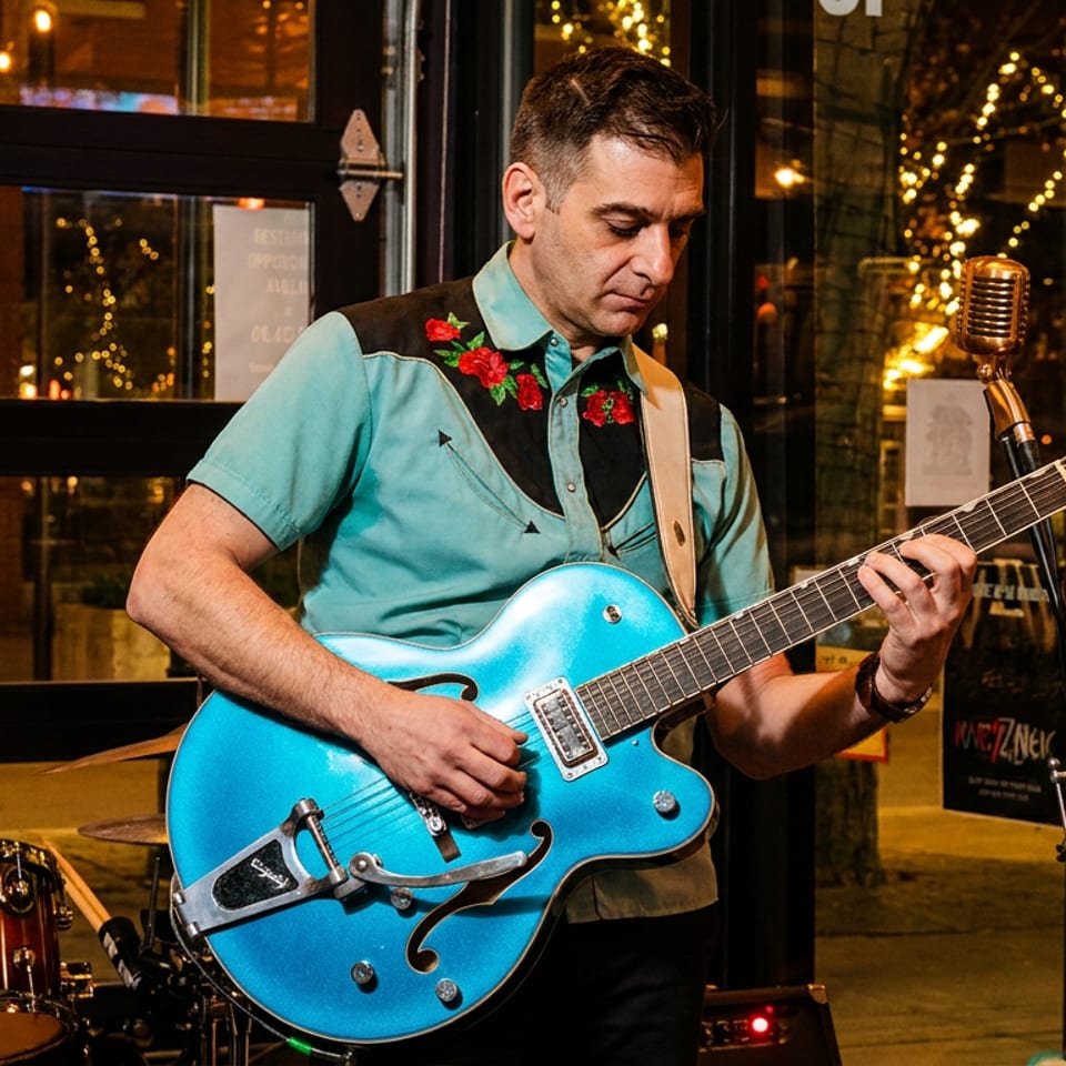 A musician in a teal shirt with red roses plays a blue electric guitar in a restaurant.