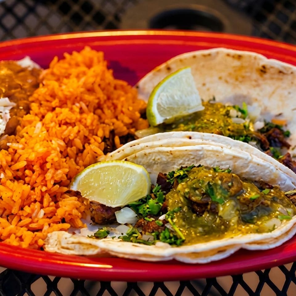 Two tacos with green salsa and lime, served with rice and refried beans on a red plate.